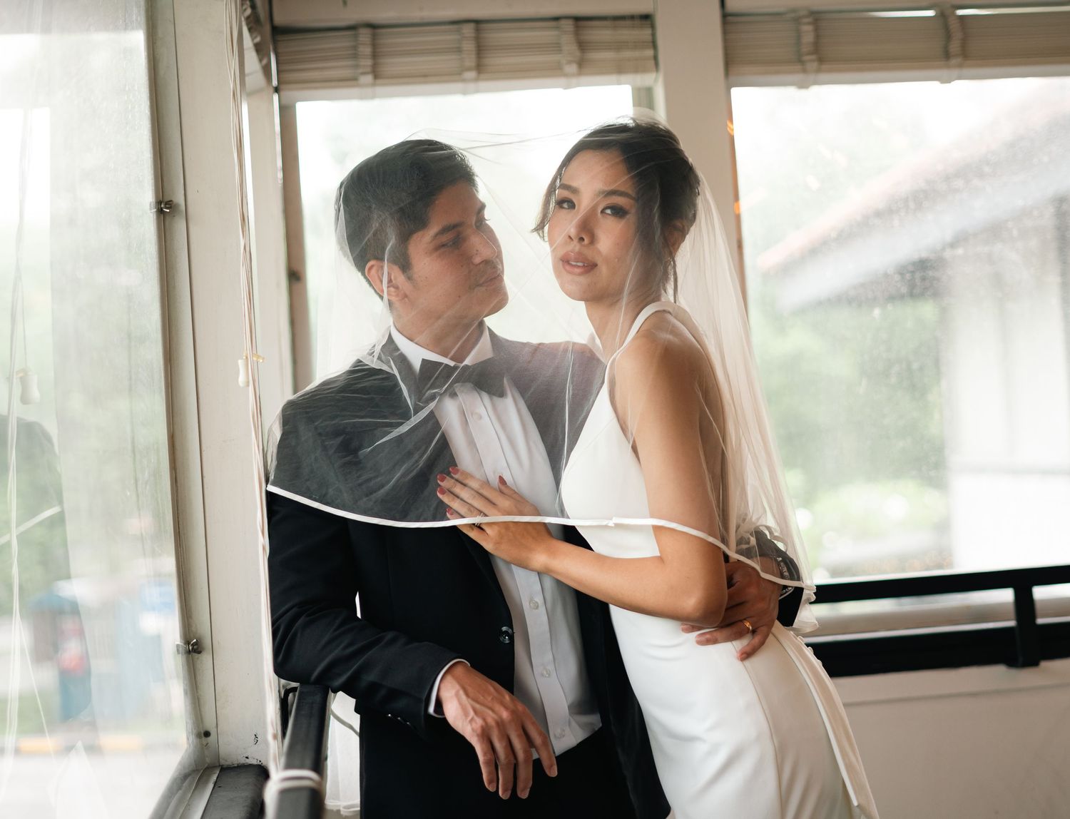 A couple pours champagne into a tower of glasses at their wedding celebration with a white curtain backdrop and floral decor.