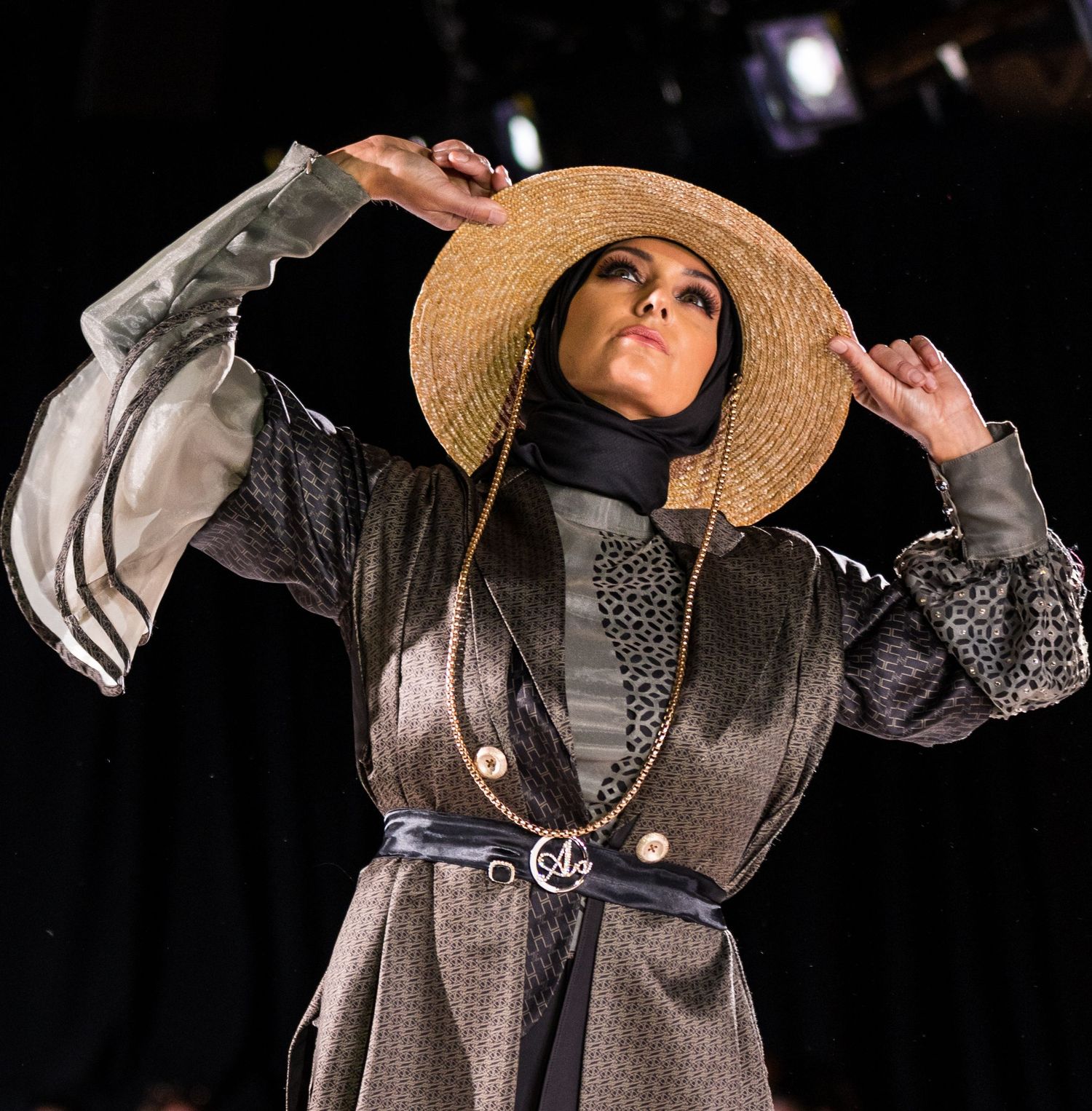 A fashion model walks the runway wearing a belted gray coat and wide-brimmed straw hat during a fashion show.