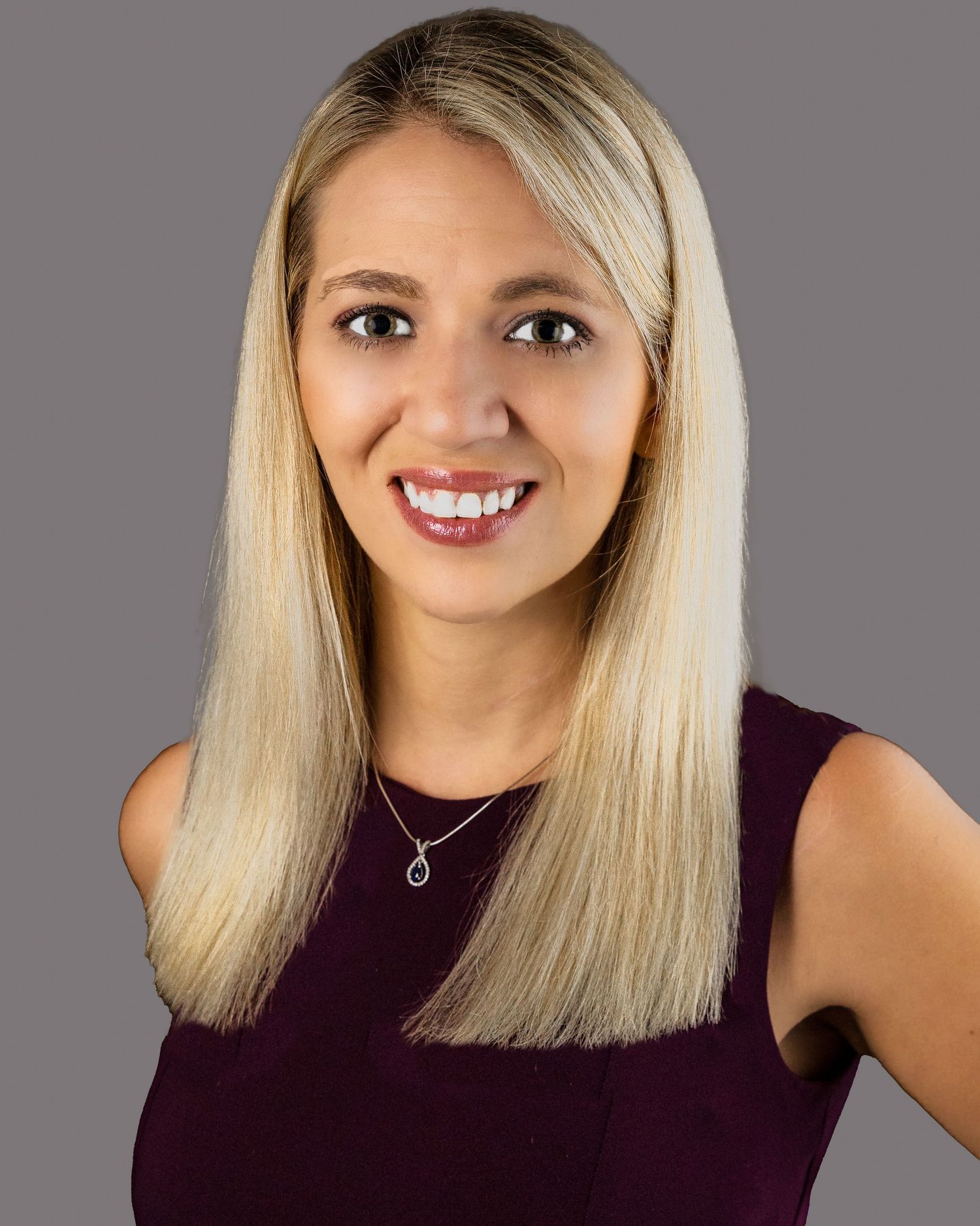 Professional headshot of a person with long blonde hair wearing a purple sleeveless top and delicate necklace against gray background.
