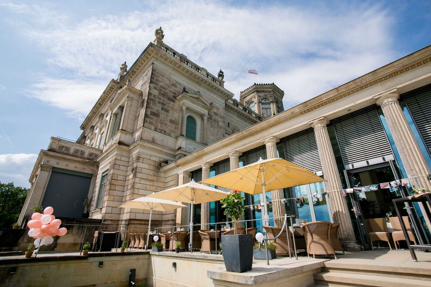 Elegant historic building with yellow umbrella on outdoor patio and decorative stone architecture against blue sky.