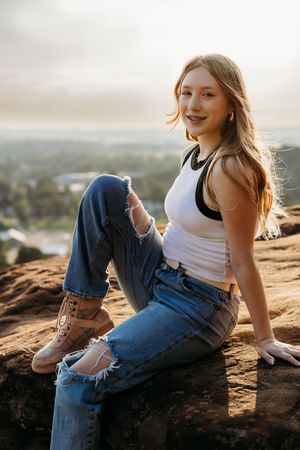 Person in ripped jeans and white tank top sits on rocky overlook during golden hour.
