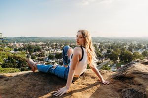 A person sits on a rocky overlook enjoying a panoramic view of a suburban town nestled among hills on a sunny day.