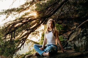 Someone sits in lotus pose meditating on a rocky cliff during golden hour, with sunlight filtering through tree branches.