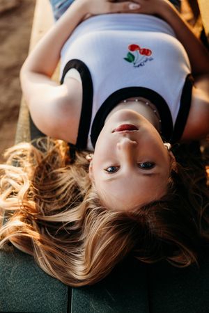 An artistic overhead portrait shot showing wavy blonde hair spread out on a green surface, wearing a white shirt with cherry design.