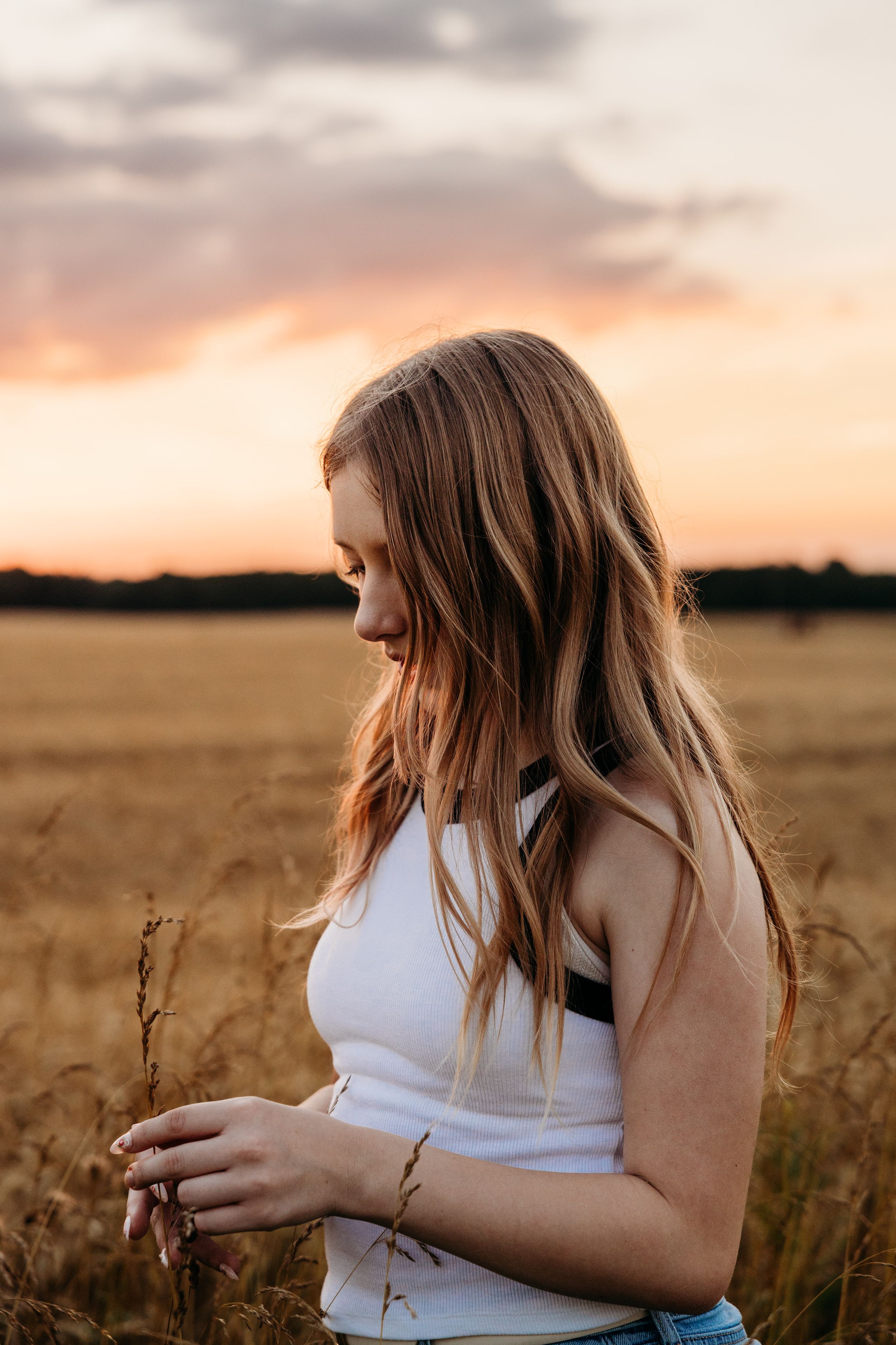 Person in white top gazes down at wheat stalks while standing in golden field during sunset.