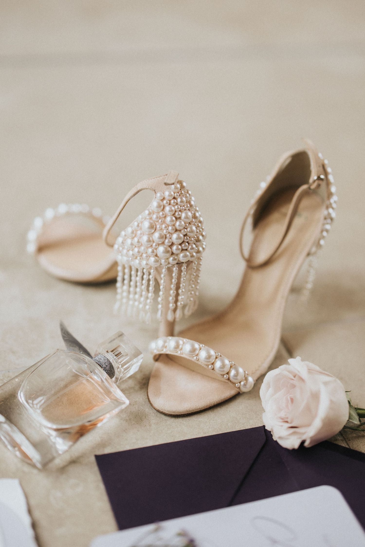 Elegant bridal shoes with pearl details displayed alongside a perfume bottle and delicate pink rose on beige background.