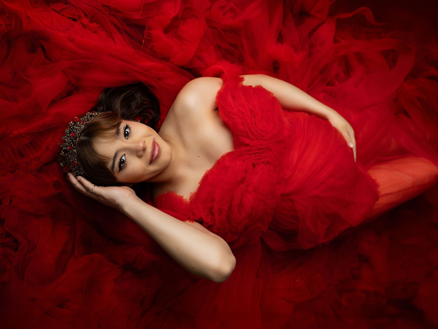 A figure lying on flowing red fabric wearing an elegant red gown and tiara in a dramatic fashion portrait.