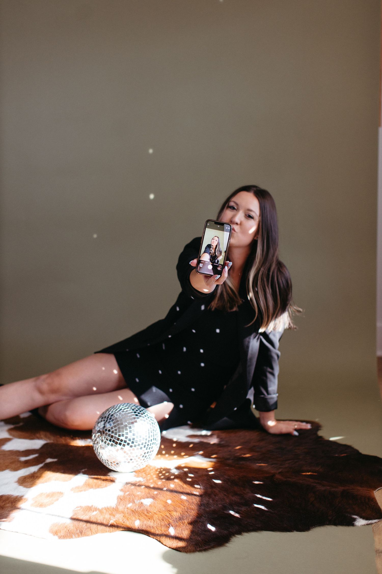 Person in black dress sitting on cowhide rug with disco ball and retro phone for stylized photoshoot.
