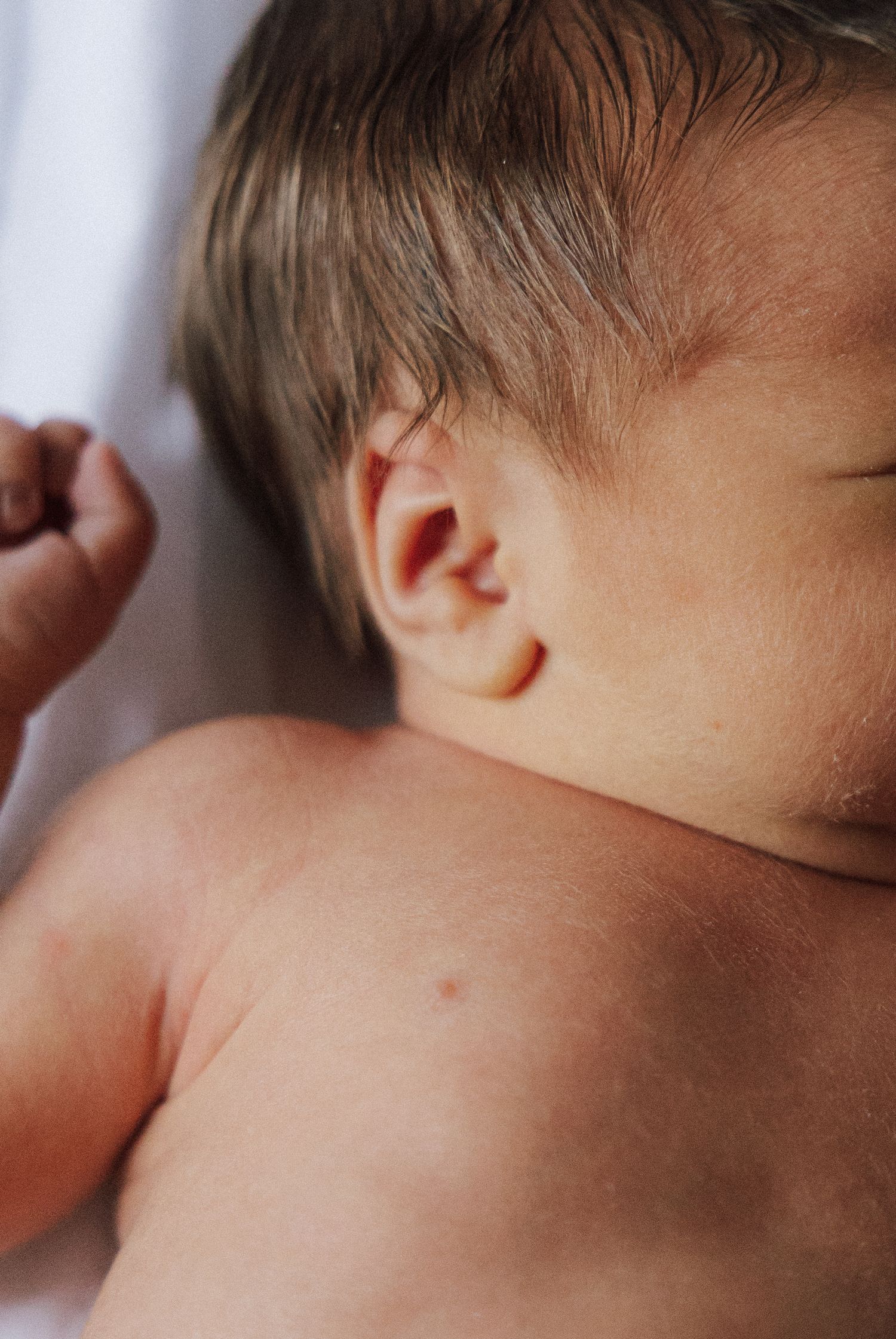 Close-up view of a sleeping baby's ear and shoulder showing delicate newborn skin in natural light.