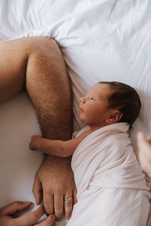 A newborn baby wrapped in white linen rests peacefully next to an adult's arm on white bedding.