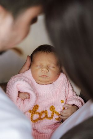 A sleeping baby wearing a pink knitted sweater with gold lettering detail lies peacefully against white bedding.