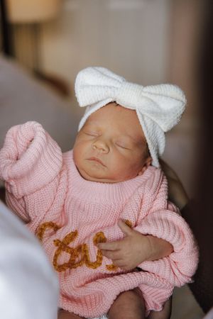 Sleeping newborn wearing a soft pink knitted sweater and white bow headband in a peaceful pose.