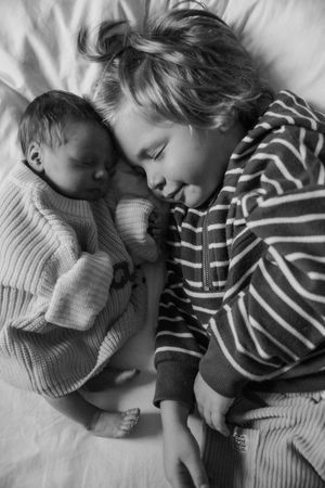 A tender black and white photo shows a newborn baby and older sibling sleeping peacefully side by side on white bedding.
