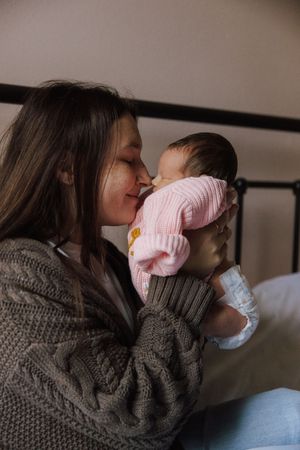Parent in a gray sweater lovingly cuddles and kisses a baby wearing pink on a couch in soft lighting.