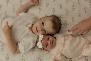 A baby lies on white bedding while laughing and playing with someone's hands reaching from outside the frame.