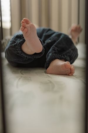 Close-up view of tiny baby feet in dark pants lying on a light floor, seen through crib bars.