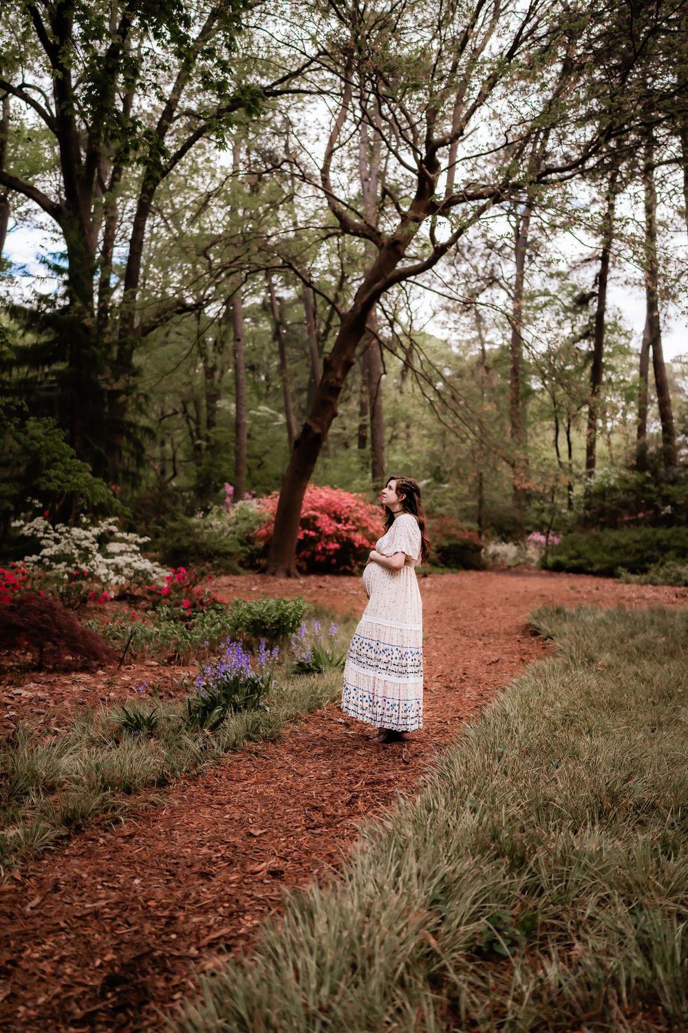 A woman in a white lace dress poses on a red dirt path surrounded by lush forest and flowering bushes.