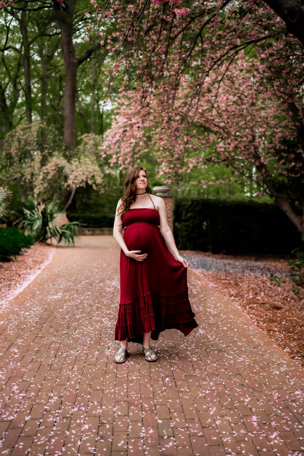 Cherry blossom petals scatter across a garden path as a figure in a burgundy dress poses beneath blooming trees.
