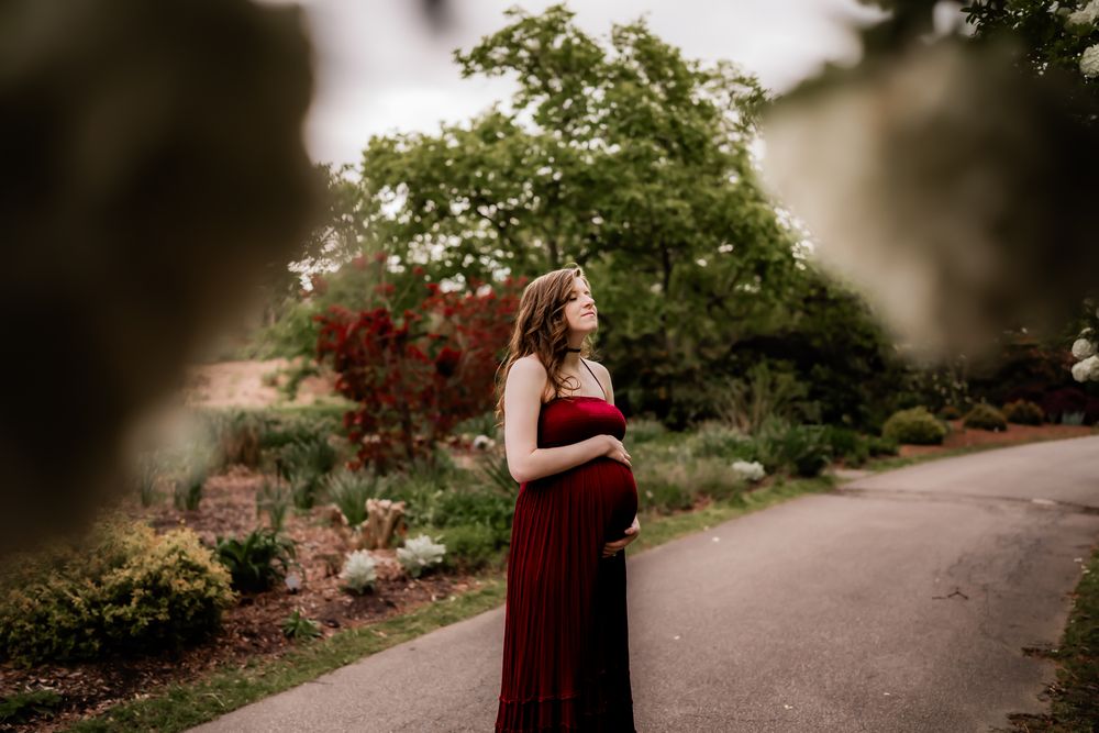 A couple in burgundy formal attire pose together during their engagement photo session in a garden setting.
