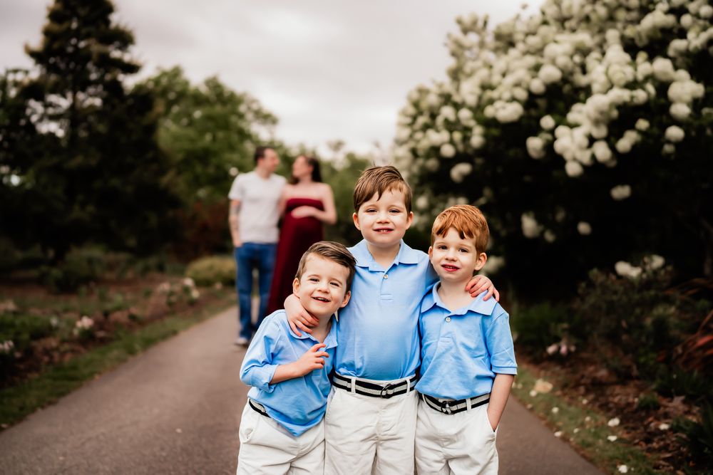 Three young boys in blue shirts and white pants smile and laugh together in a garden setting.