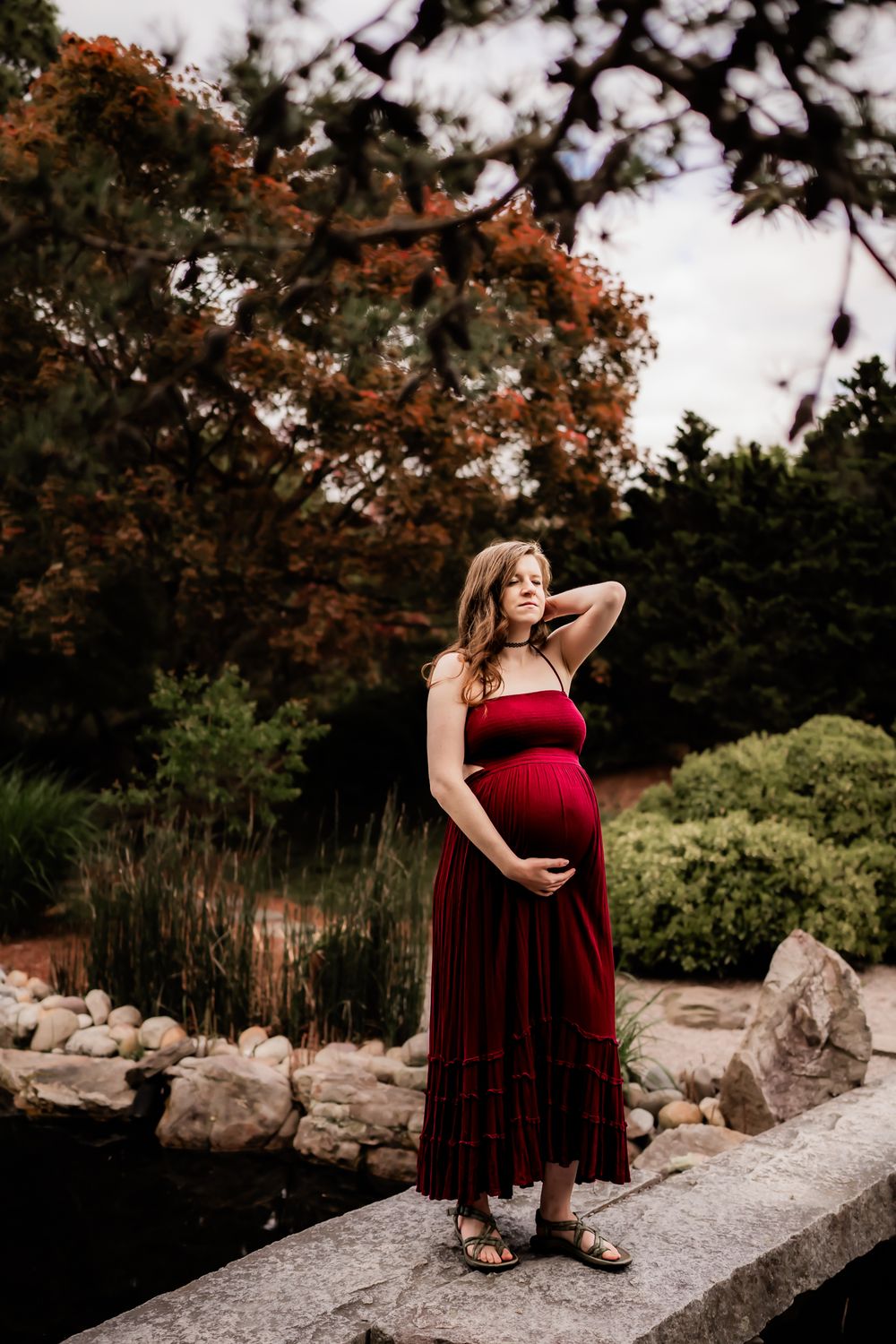 A pregnant woman in a burgundy velvet dress poses gracefully on a stone wall near autumn trees.