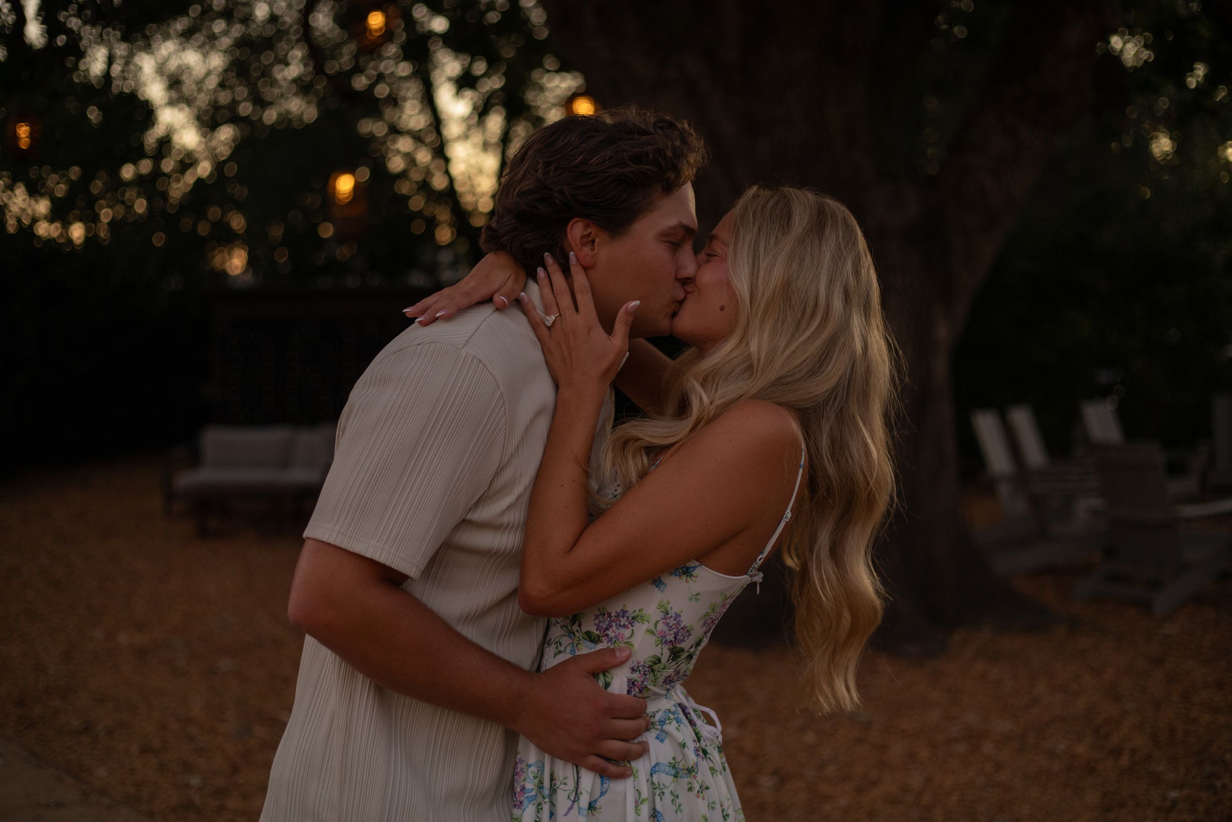 Silhouetted against a glowing sunset, a couple in casual attire shares an intimate moment in a park setting.