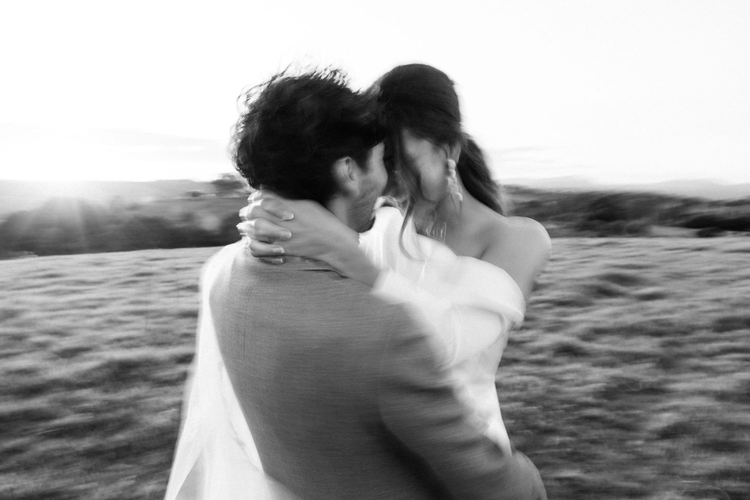 An intimate black and white photo of a couple embracing at sunset on a hilltop.