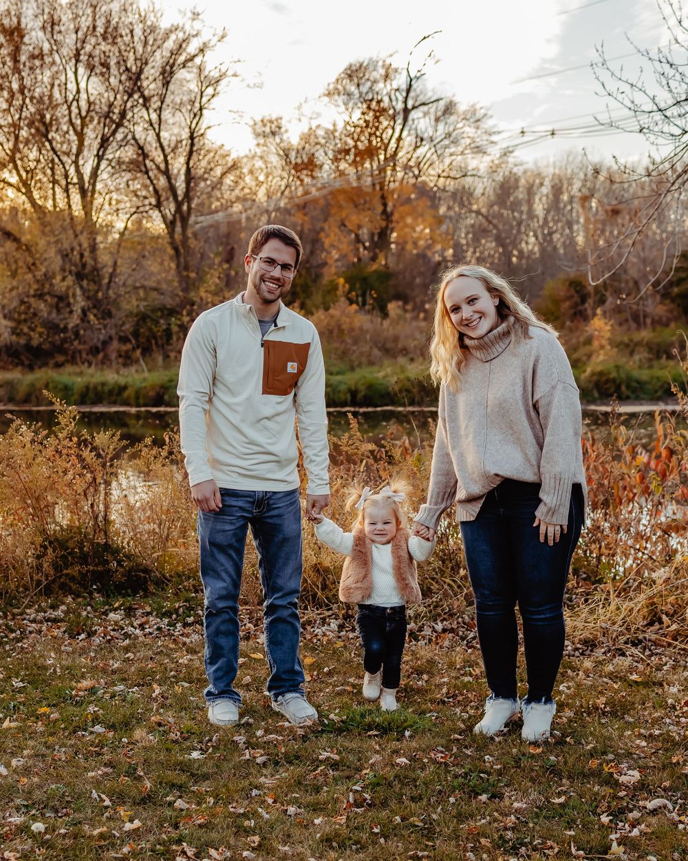 A family of three poses together in an autumn setting with golden foliage and warm sunset lighting in the background.