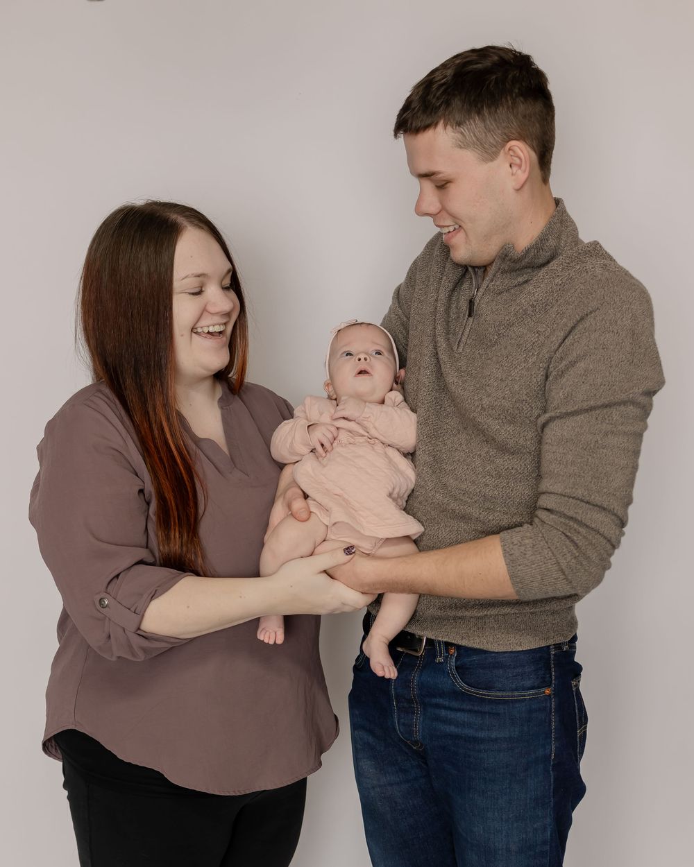A baby wearing a pink onesie is held by parents against a light gray backdrop for a family portrait.