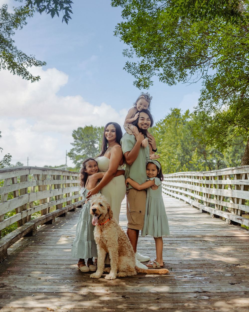 Family and their furry friend pose together on a wooden bridge surrounded by lush greenery on a sunny day.