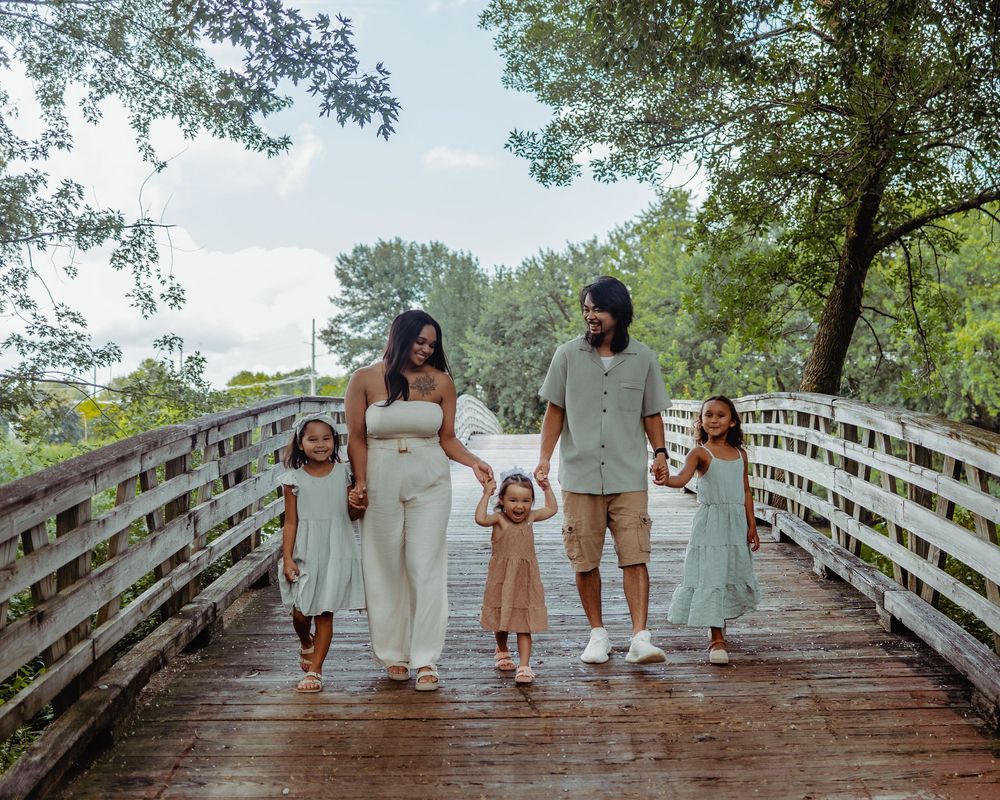 A loving family walks together holding hands on a wooden bridge surrounded by green trees on a summer day.
