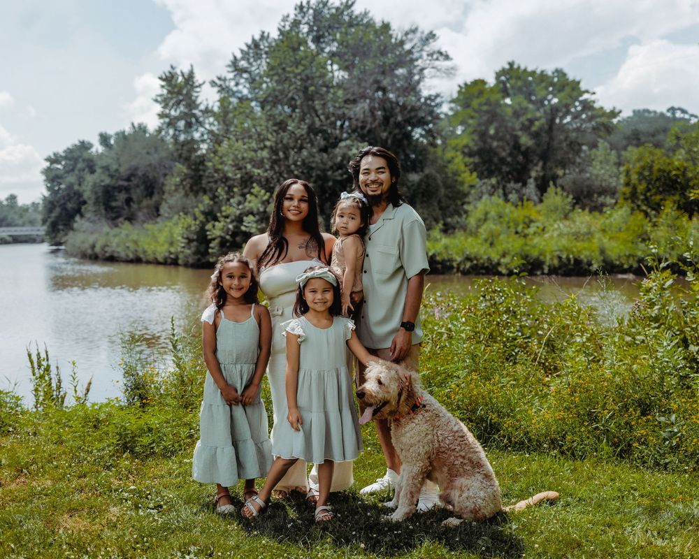 A family in matching blue outfits poses together with their dog by a scenic lake surrounded by summer wildflowers.