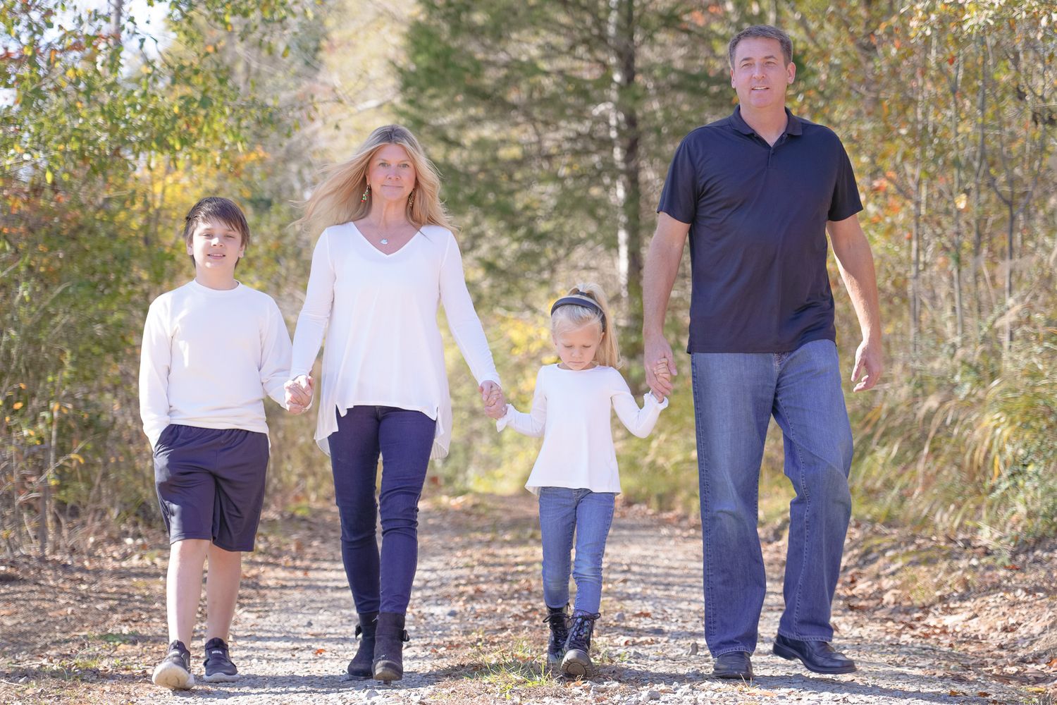 A family walks together holding hands on a peaceful nature trail surrounded by trees and autumn foliage.