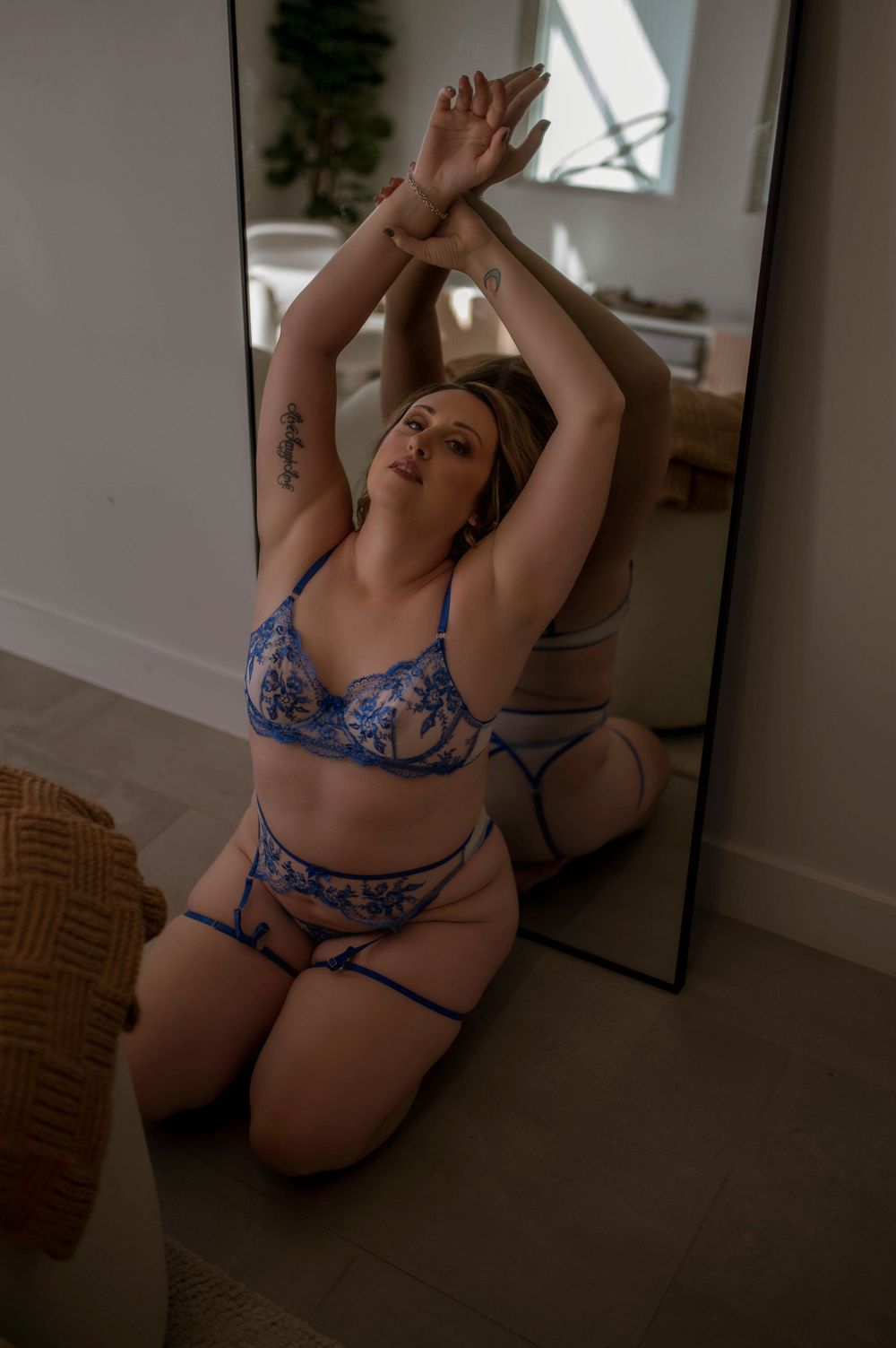 Woman in blue lace lingerie set kneeling by a mirror with arms raised, showcasing elegant embroidered bra and garter.