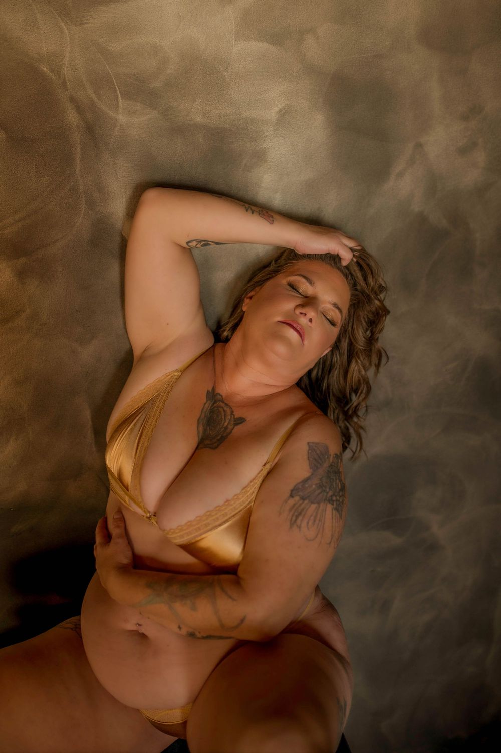 A tattooed plus-size woman poses confidently in gold lingerie against a smoky dark background.