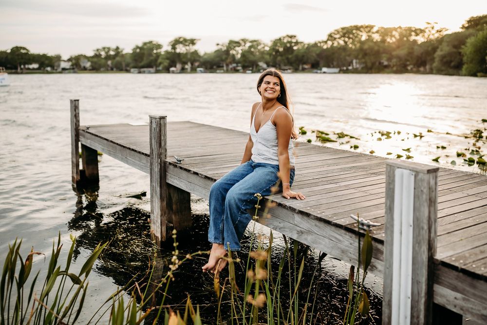 Person sits on wooden dock overlooking tranquil lake at sunset with boats and palm trees in background.