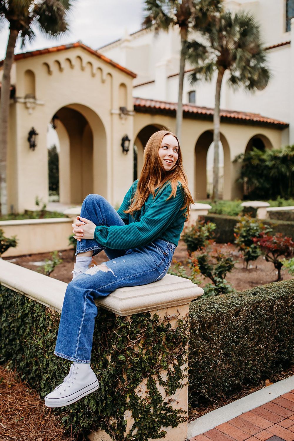 A woman in blue jeans and white sneakers sits casually on a low wall near Mediterranean-style architecture.