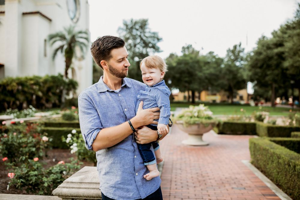 A loving parent in a blue shirt holding and playing with a toddler outdoors in a garden setting.