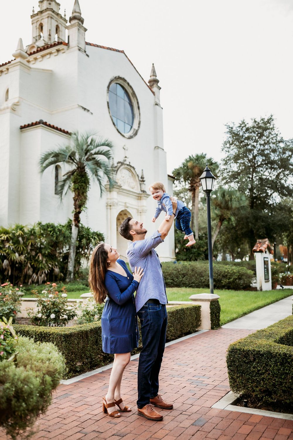 A couple dances and twirls in front of a white Spanish-style church building on a brick path surrounded by palm trees.