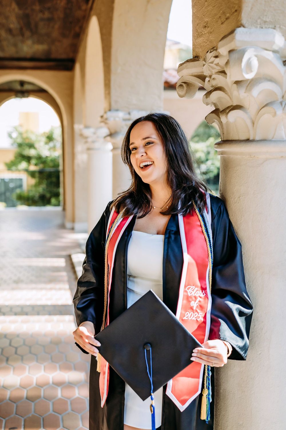 Graduate poses in graduation regalia with cap and sash under a colonnade of arches during photoshoot.