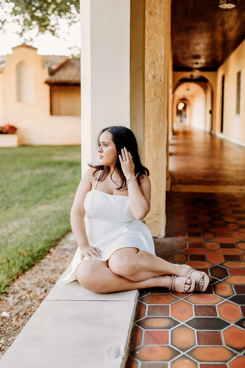 Person in white dress poses elegantly along a covered walkway with terracotta tiles and columns.