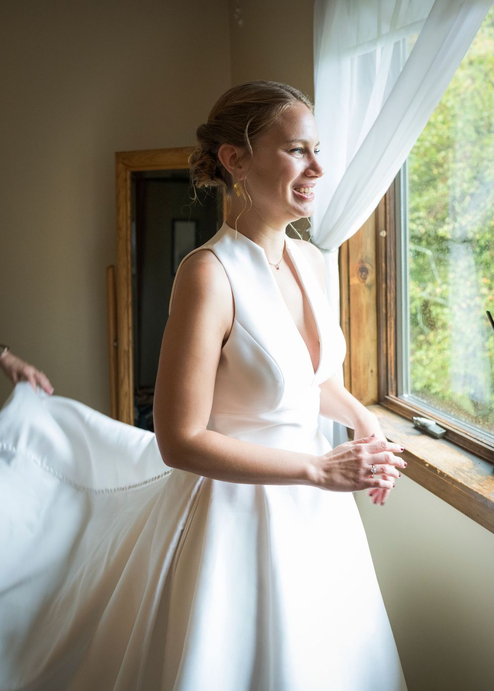 A person in an elegant white wedding dress gazes thoughtfully out a sunlit window.