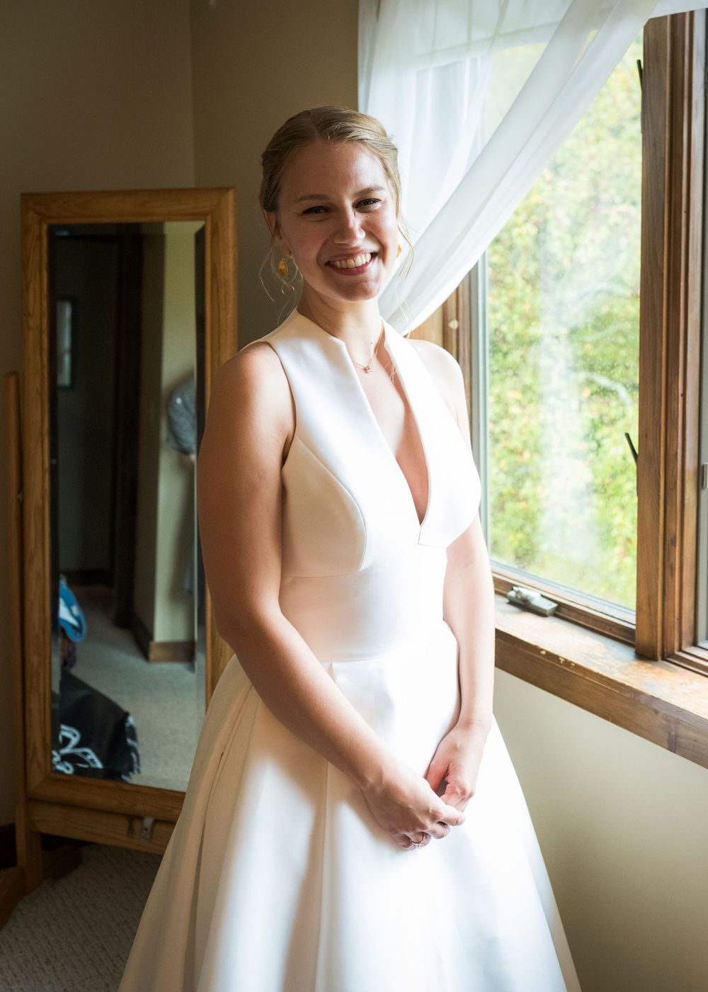 A bride in an elegant white V-neck wedding dress stands by a window with natural light streaming in.