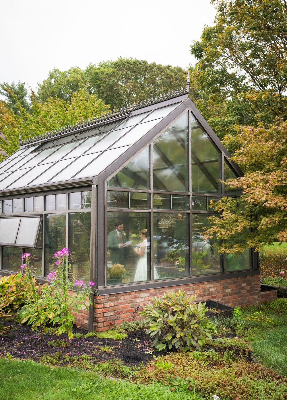 A modern glass greenhouse with brick foundation surrounded by lush garden landscaping and autumn trees.