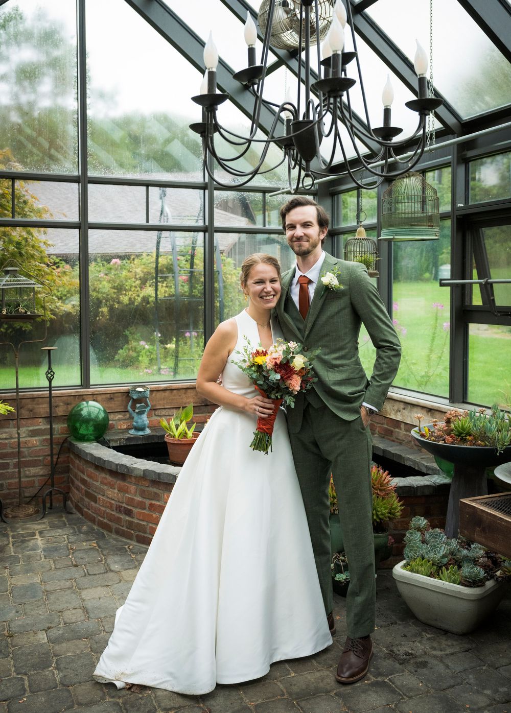 Wedding couple posing in a greenhouse with glass walls and potted plants surrounding them.