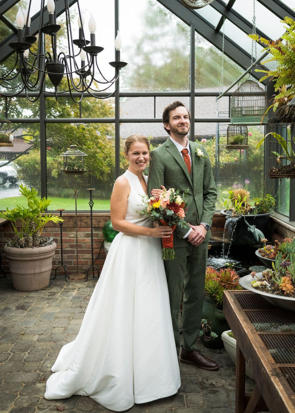 Wedding couple poses in a greenhouse setting with natural light streaming through glass panels.