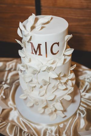Elegant three-tier white wedding cake decorated with delicate sugar flowers and monogram M|C on satin tablecloth.