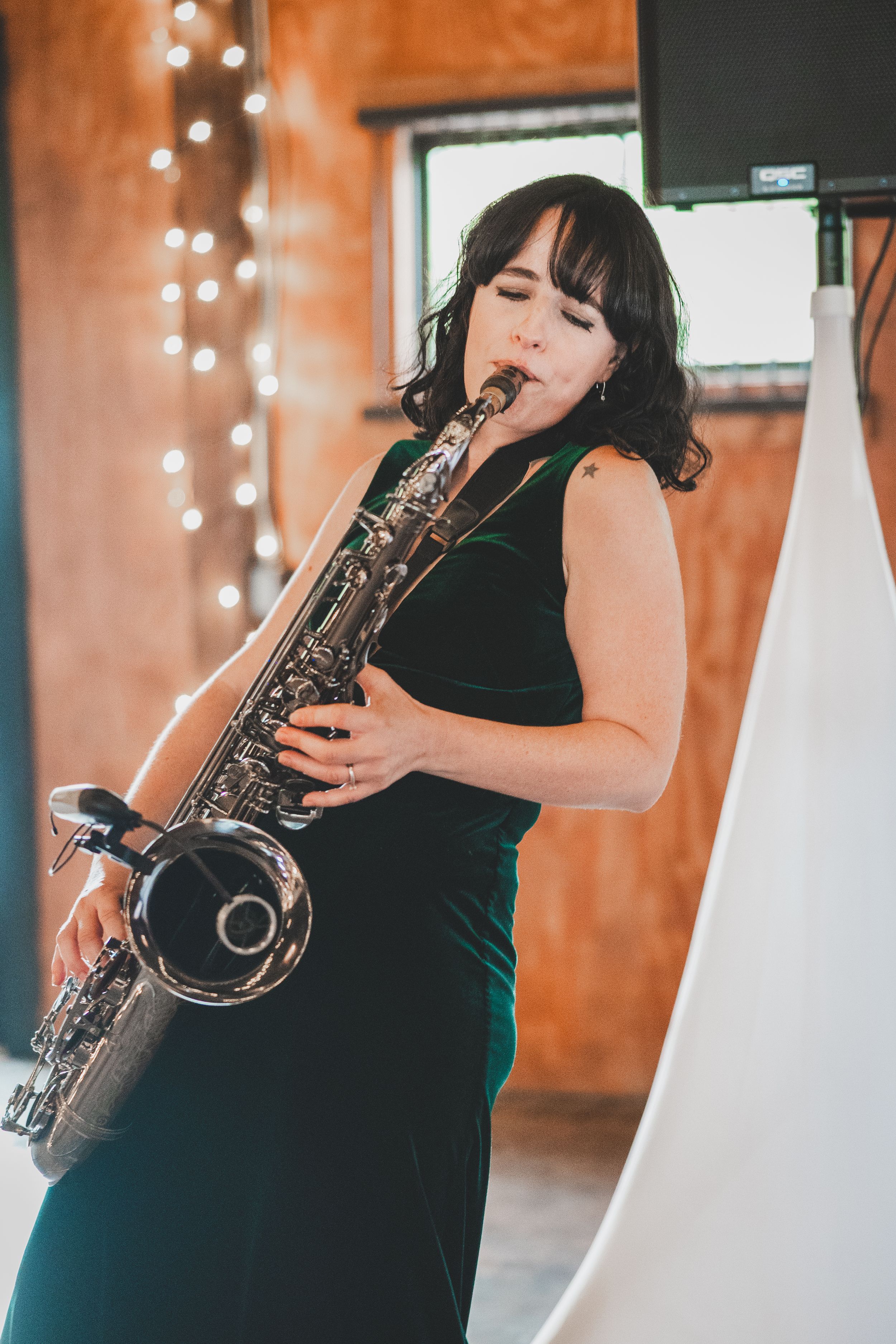 A musician in a black dress plays saxophone on stage with string lights in the background.