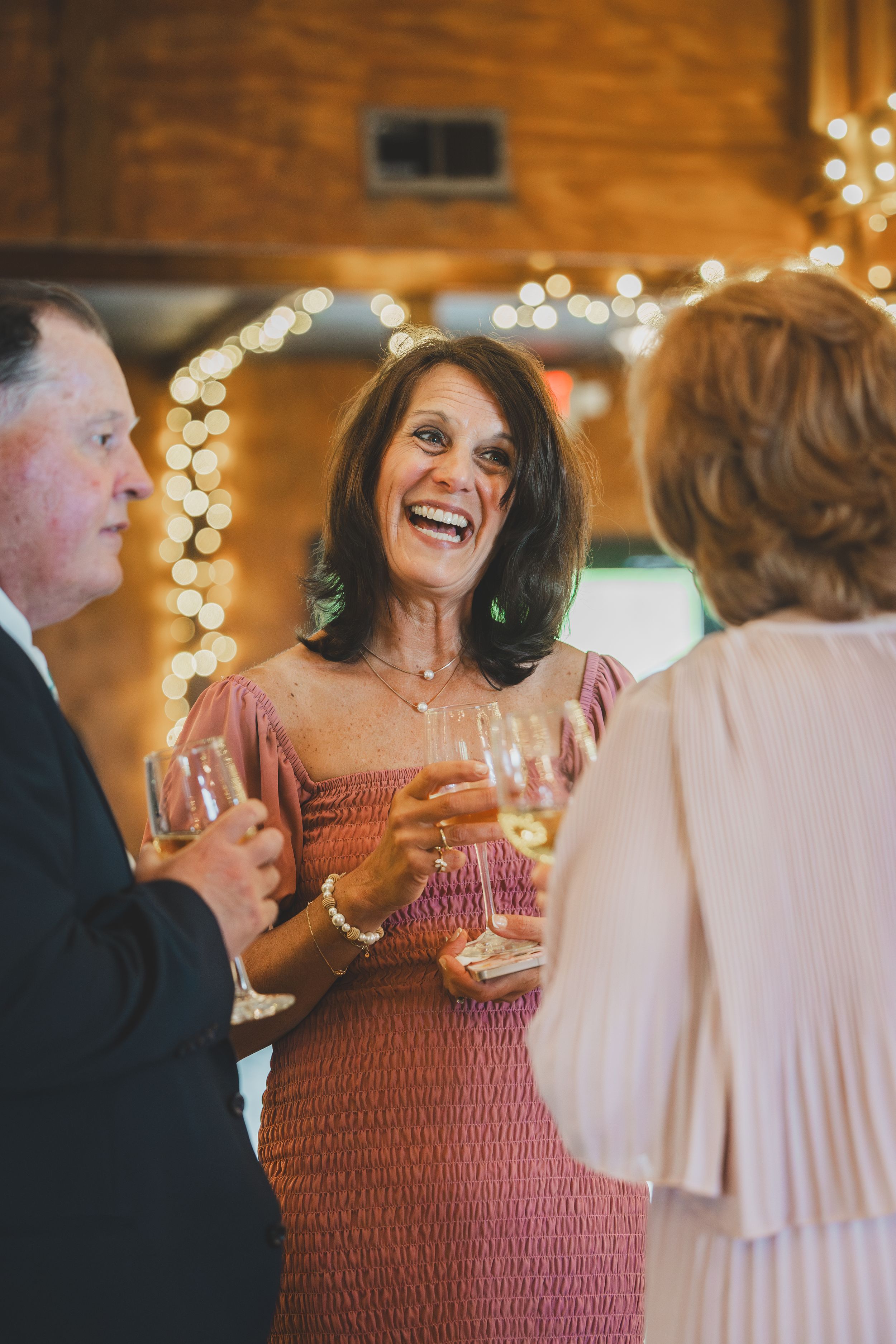 A person in a red smocked dress laughs while holding a wine glass during a festive indoor event with string lights.