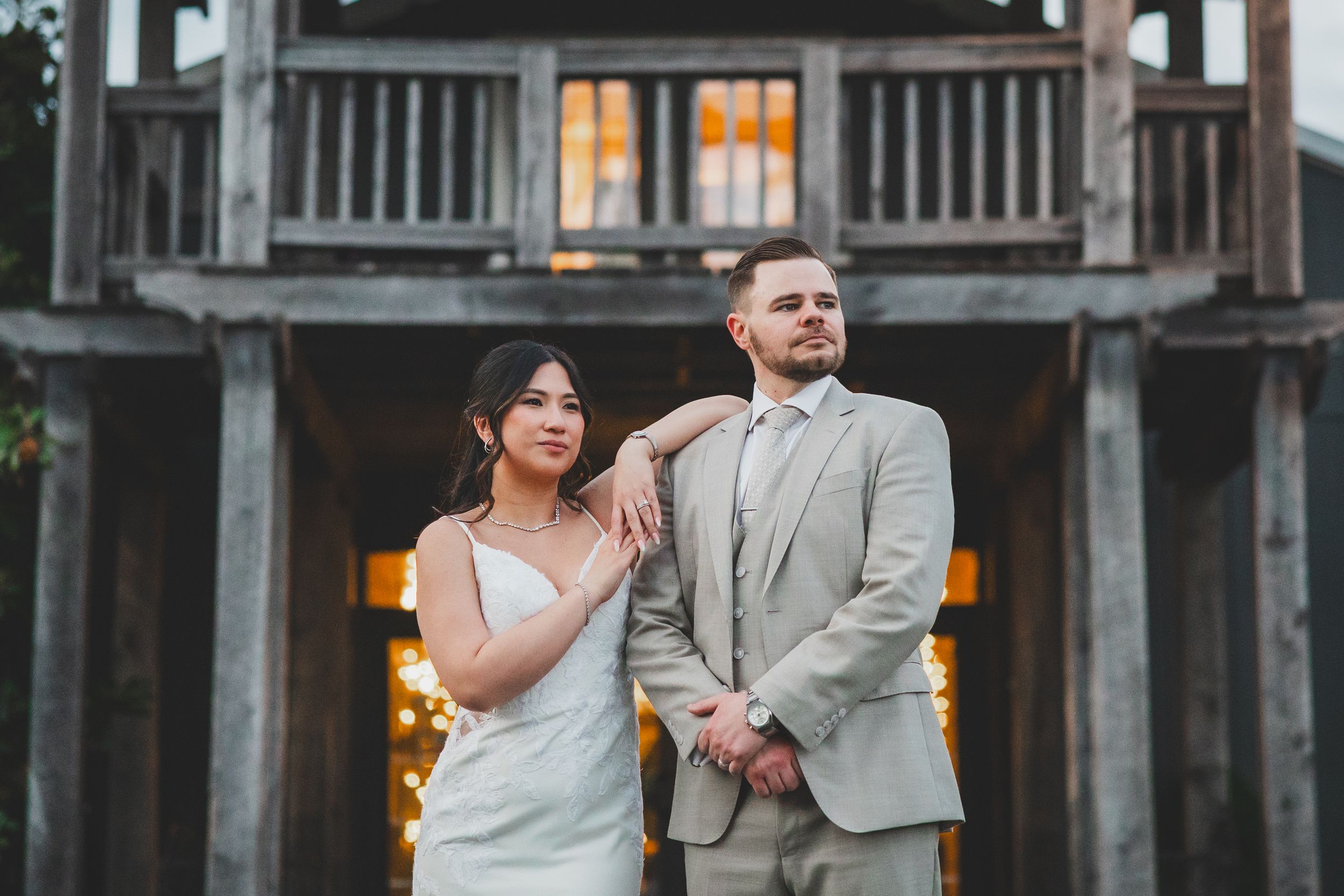 A couple in wedding attire poses together in front of a rustic wooden building with glowing lights at dusk.